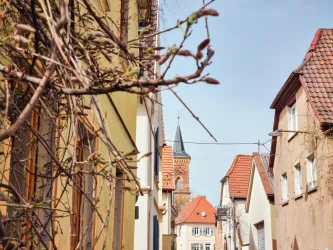 Eine malerische Gasse mit bunten Häusern und einem Kirchturm im Hintergrund. Die Pflanzen im Vordergrund verleihen der Szene einen charmanten Charakter.