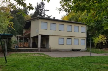 Ein zweigeschossiges Gebäude in einer grünen Umgebung. Es hat gelbe Wände und mehrere Fenster, die mit Rollläden versehen sind.