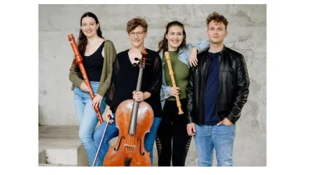 Eine Gruppe von vier Musikern steht zusammen und hält verschiedene Instrumente. Sie lächeln und posieren vor einem schlichten Hintergrund.