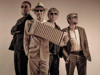 Vier Frauen in eleganten Kleidern sitzen um ein Klavier. Sie lächeln und posieren zusammen für das Foto.