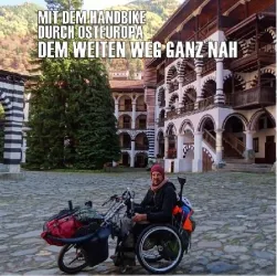 Ein Mann sitzt auf einem Handbike in einem malerischen Innenhof mit traditionellen Gebäuden. Im Hintergrund sind grüne Bäume und Berge zu sehen.
