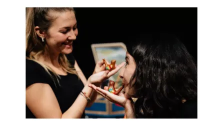 Zwei Frauen lachen und spielen mit kleinen Figuren in einer dunklen Umgebung. Eine Frau hält eine Figur in der Hand, während die andere sie neugierig anschaut.