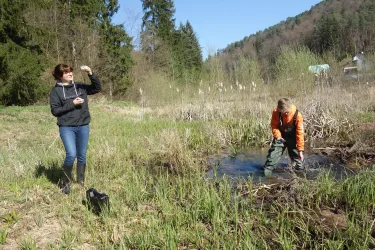 Eine Frau beobachtet einen Jungen, der in einem feuchten Gebiet spielt. Im Hintergrund sind Bäume und ein blauer Himmel zu sehen.