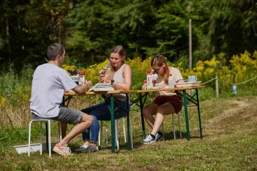 Drei Personen sitzen an einem Tisch im Freien und genießen ihre Mahlzeiten. Im Hintergrund sind grüne Wiesen und Bäume zu sehen.