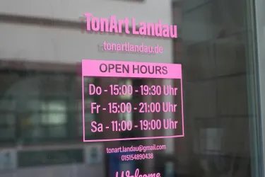 Ein geöffnetes Schild mit den Öffnungszeiten von TonArt Landau. Die Zeiten sind deutlich sichtbar in pinker Schrift.
