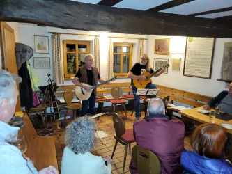 Zwei Musiker spielen Gitarre in einem gemütlichen Raum. Eine kleine Menge von Zuhörern hört aufmerksam zu.