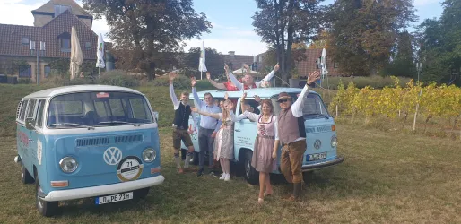 Eine Gruppe von Menschen steht fröhlich neben zwei blauen Volkswagen Bullis am Deutschen Weintor. Im Hintergrund sind Bäume und Weinreben zu sehen.