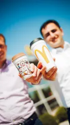 Zwei Männer halten jeweils einen Kaffeebecher in der Hand. Im Hintergrund ist ein klarer Himmel und ein Teil eines McDonald's-Restaurants zu sehen.