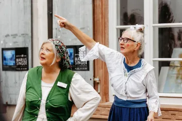 Zwei Frauen in traditionellen Kleidern stehen im Freien. Eine von ihnen zeigt auf etwas, während die andere aufmerksam zuschaut.