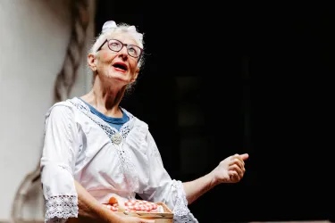 Eine ältere Frau mit Brille trägt ein traditionelles Outfit und sieht fröhlich aus. Sie hält eine Schachtel in der Hand und scheint etwas zu erzählen.