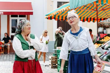 Zwei Frauen in traditionellen Kostümen stehen auf einem Markt und lachen. Im Hintergrund sind Stände mit frischem Obst und Gemüse zu sehen.