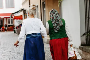 Eine ältere Frau mit grauen Haaren und einer Brille schaut nach oben. Im Hintergrund sind Holzfenster und ein Tisch mit Blumen zu sehen.
