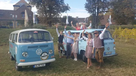Eine Gruppe von fröhlichen Menschen in traditioneller Tracht steht neben zwei blauen VW-Bussen. Sie winken in die Kamera vor einer malerischen Kulisse.