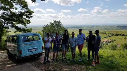 Eine Gruppe von Menschen steht lächelnd im Freien. Im Hintergrund ist eine weite Landschaft mit Wiesen und Bäumen zu sehen, sowie ein blaues Fahrzeug.