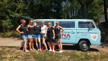 Sechs Frauen stehen fröhlich neben einem blauen VW-Bus. Sie tragen Sommerkleidung und genießen einen sonnigen Tag im Freien.