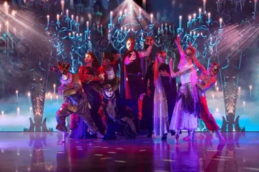 Eine dramatische Aufführung mit Tänzern in bunten Kostümen und Masken. Im Hintergrund leuchtet eine geheimnisvolle Kulisse mit Kerzen und Schatten.