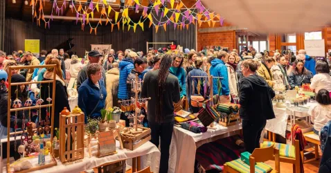 Ein belebter Markt mit vielen Menschen, die verschiedene Stände besuchen. Farbenfrohe Dekorationen hängen von der Decke und schaffen eine festliche Atmosphäre.