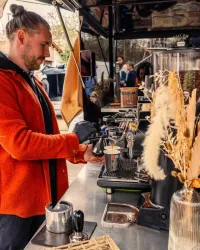 Ein Barista bereitet Kaffee an einem Foodtruck. Die Umgebung ist lebhaft mit Menschen und dekorativen Pflanzen.