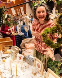 Eine lächelnde Frau steht an einem Verkaufsstand mit vielen handgefertigten Dekorationen. Um sie herum hängen grüne Pflanzen und die Tische sind mit verschiedenen Kunstwerken geschmückt.