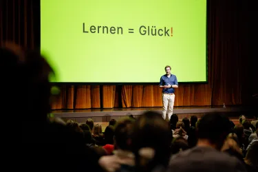Ein Redner steht auf einer Bühne vor einem großen, grünen Bildschirm mit der Aufschrift „Lernen = Glück!“. Im Publikum sitzen viele Menschen und hören aufmerksam zu.