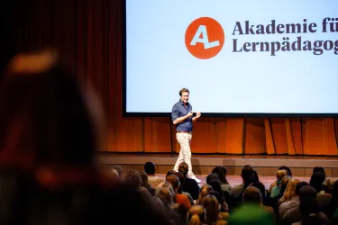 Ein Redner steht auf der Bühne vor einem Publikum. Im Hintergrund ist das Logo der Akademie für Lernpädagogik zu sehen.