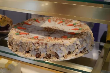 Eine große Karottenkuchen auf einem Glasregal. Der Kuchen ist mit kleinen Karotten und Mandeln dekoriert.