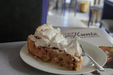 Ein Stück Kuchen mit einer dicken Schicht Sahne auf einem weißen Teller. Im Hintergrund ist eine unwirkliche Café-Atmosphäre sichtbar.