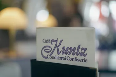Ein Schild mit der Aufschrift "Café Kuntz" und dem Zusatz "Conditorei Confiserie". Der Hintergrund zeigt eine gemütliche Café-Atmosphäre.