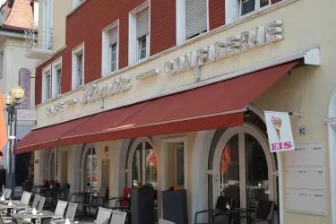 Ein gemütliches Café mit einem roten Markisen und einem Schild für Eis. Die Fassade ist hell und einladend, mit mehreren Stühlen im Freien.