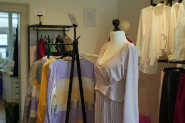 Ein modisches Geschäft mit einer Schneiderpuppe und verschiedenen Kleidungsstücken auf Kleiderständern. Die Farbauswahl reicht von sanften Pastelltönen bis zu kräftigen Farben.