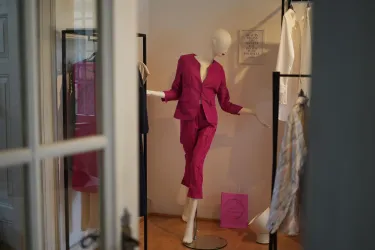 Eine Schaufensterpuppe trägt einen leuchtend pinken Anzug. Im Hintergrund sind weitere Kleidungsstücke auf Ständern zu sehen.
