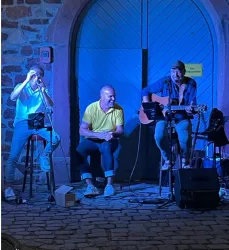Drei Musiker stehen vor einer blauen Tür und spielen live Musik. Es ist eine fröhliche Atmosphäre mit buntem Licht.