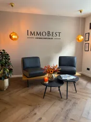 Ein modernes Wartezimmer mit zwei schwarzen Sesseln und einem kleinen Tisch. An der Wand steht "ImmoBest" und es gibt eine Blumenarrangement sowie warme Lichtquellen.