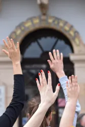 Hochgestreckte Hände vor einem Gebäude mit einer markanten Schrift. Die Hände zeigen verschiedene Nagellackfarben.