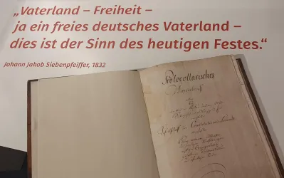 Ein historisches Buch mit handschriftlichem Inhalt und einer bedeutenden zitierten Aussage über Freiheit und das Vaterland. Im Hintergrund steht der Name Johann Jakob Siebenpfiffer aus dem Jahr 1832.