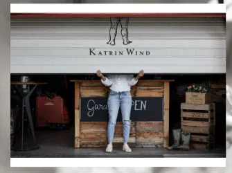Eine Person hebt eine Rolltür mit dem Schriftzug „Katrin Wind“ darüber. Im Hintergrund sind Holzpaletten und ein Schild mit der Aufschrift „Geriß OPEN“ zu sehen.