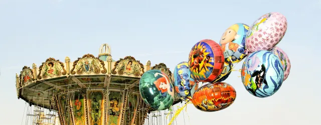 Ein Jahrmarkt mit einem Karussell im Hintergrund und bunten Ballons, die Charaktere aus Kinderfilmen zeigen. Die Atmosphäre ist fröhlich und einladend.