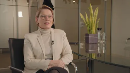 Eine Frau sitzt in einem modernen Büro und lächelt. Im Hintergrund sind Pflanzen und ein Glastisch zu sehen.