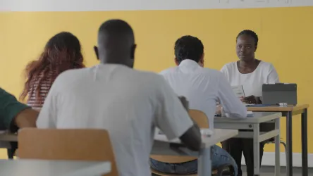 Eine Lehrerin spricht mit Schülern in einem Klassenzimmer. Die Wände sind gelb gestrichen und die Schüler sitzen an einfachen Tisch.