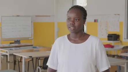 Eine Frau steht in einem Klassenzimmer und spricht. Im Hintergrund sind Tische und eine Tafel sichtbar.