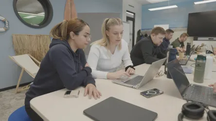 Zwei Frauen arbeiten konzentriert an einem Laptop in einem modernen Büro. Im Hintergrund sind weitere Personen an ihren Laptops beschäftigt.