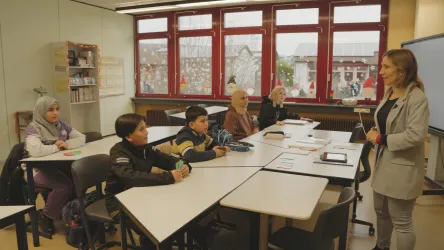 Eine Lehrerin steht vor einer Gruppe von Schülern in einem Klassenzimmer. Die Schüler hören aufmerksam zu und sind an einem Tisch versammelt.