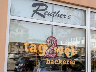 Ein freundliches Bäckerei-Schild mit dem Namen "Reuther's tag2wei backerei". Die Fenster reflektieren die Umgebung und laden Passanten ein.