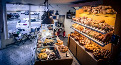 Eine gemütliche Bäckerei mit frisch gebackenem Brot und Gebäck auf Regalen. Am Fenster stehen kleine Tische mit Stühlen und es ist hell erleuchtet.