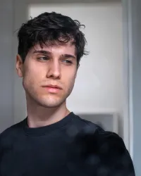 Ein junger Mann mit dunklen, lockigen Haaren blickt nachdenklich zur Seite. Er trägt ein einfaches schwarzes Shirt und der Hintergrund ist unscharf.