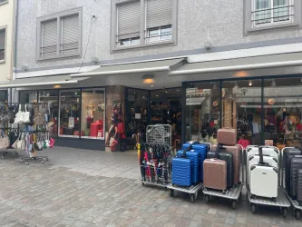 Ein Geschäft mit einer Auswahl an Taschen und Koffern steht an einer gepflasterten Straße. Vor der Ladentür sind mehrere Koffer ausgestellt.
