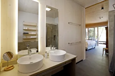 Ein modernes Badezimmer mit zwei Waschbecken und großflächigem Spiegel. Im Hintergrund ist ein gemütliches Schlafzimmer mit großen Fenstern zu sehen.