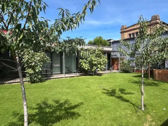 Ein Garten mit grünem Rasen und kleinen Bäumen. Im Hintergrund sind moderne Gebäude und eine klare, blaue Himmel zu sehen.