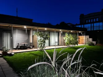 Ein modernes Haus mit einer Holzt terrasse, umgeben von Grünpflanzen. Es ist Nacht und die Umgebung ist sanft beleuchtet.