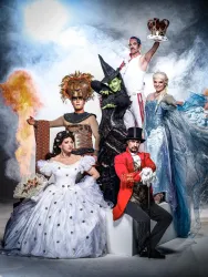 Eine Gruppe von Menschen in festlichen Kostümen steht vor einem dramatischen Hintergrund. Die Charaktere scheinen aus verschiedenen berühmten Geschichten oder Musicals zu stammen.